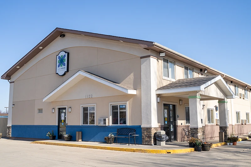 ShareHouse Grand Forks Residential two-story building with beige siding, a blue foundation stripe, and a welcoming entrance with a green trim porch.