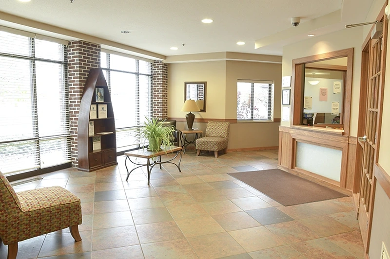 ShareHouse Fargo Residential treatment lobby featuring a colorful chair, a coffee table with a plant, a dark wood bookshelf, and a service desk, with large windows letting in natural light.
