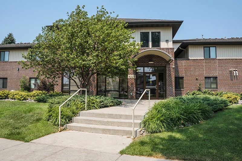 ShareHouse Fargo Residential Campus Exterior front entrance with large windows, featuring green landscaping, including shrubs and a tree.