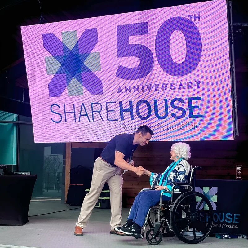 ShareHouse president Jeremy Traen shaking hands with elderly woman on stage during 50th anniversary event