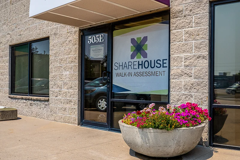Exterior view of the ShareHouse Walk-In Assessment building entrance, featuring a sign for "WALK-IN ASSESSMENT" and a round planter with colorful flowers.