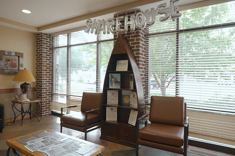 ShareHouse Fargo Residential interior cozy waiting area with two brown leather chairs, a boat-shaped bookshelf filled with brochures, and "SHAREHOUSE" spelled in metallic balloons, illuminated by natural light from large windows.