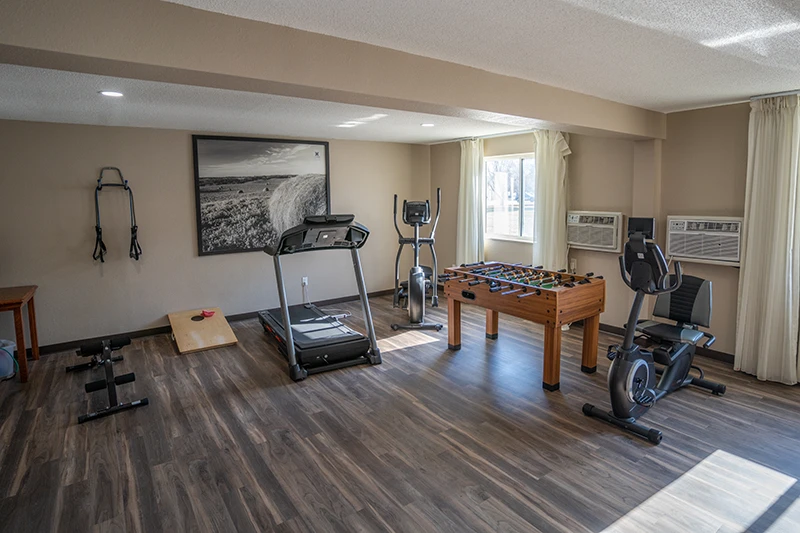 A fitness room with a treadmill, stationary bike, foosball table, dumbbells, and a cornhole board, featuring large windows, wooden flooring, and a landscape photo on the wall at ShareHouse Grand Forks Residential.