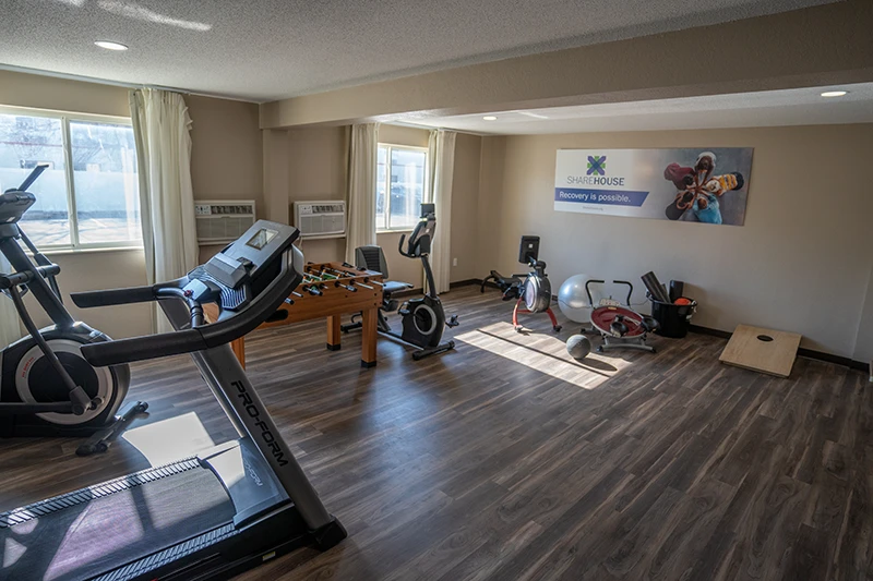 Fitness room featuring fitness equipment including a treadmill, elliptical, stationery bike, and more on wooden flooring with air conditioning and large windows at ShareHouse Grand Forks Residential.