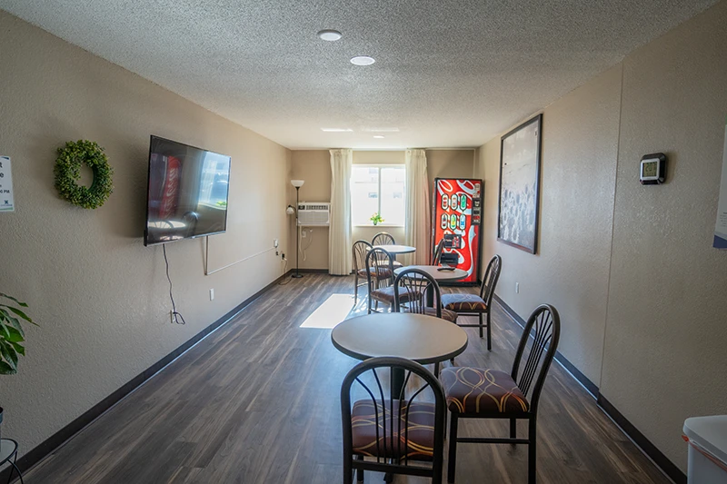 ShareHouse Grand Forks Residential common area featuring light brown walls, a wooden floor, a television, four round tables with dark-framed chairs, a vending machine, and an air conditioning unit next to a window with sheer curtains.