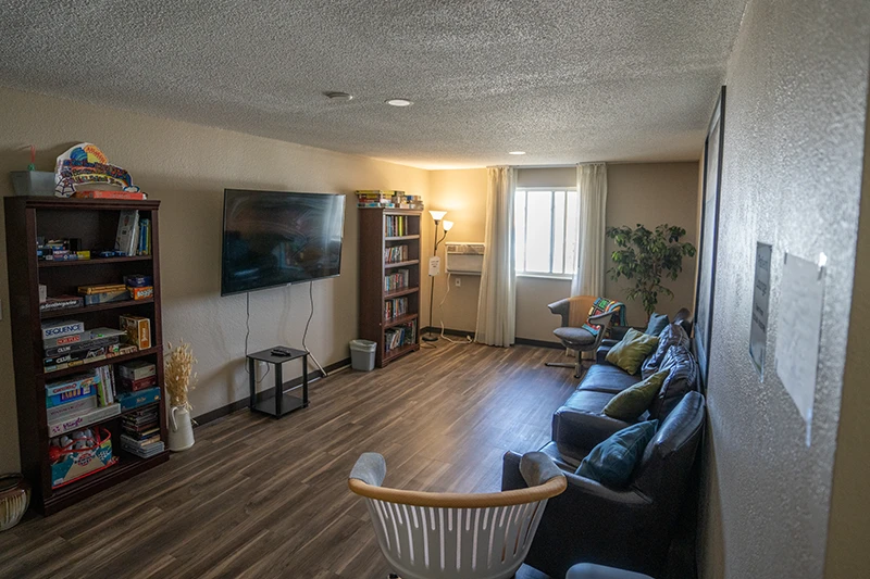 ShareHouse Grand Forks common space featuring a black couch with colorful pillows, a TV on the wall, a wooden bookshelf, and large windows with sheer curtains.