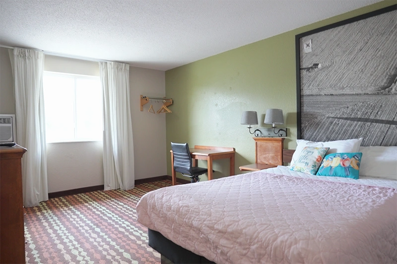 Bedroom interior featuring a made bed with a pink comforter, pillows, a large painting, wall lighting, a desk, curtains, and a dresser.