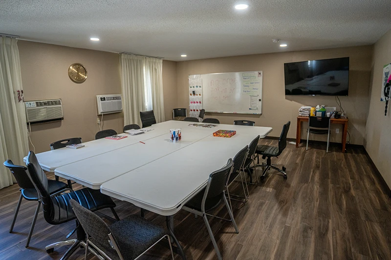 Craft room with multiple white tables placed together, black chairs, a wall clock, and a whiteboard filled with notes. There is an air conditioning unit, curtains, a flat-screen TV, and a wooden table with supplies.