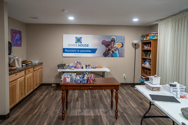 Craft room featuring tables with art supplies, a banner saying "Recovery is possible," and a bookshelf filled with items.