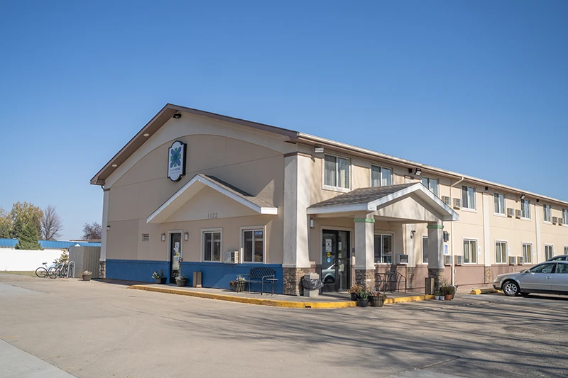 ShareHouse Grand Forks Residential building with a covered entrance under a blue sky, featuring a bicycle rack and a parked car in the foreground.