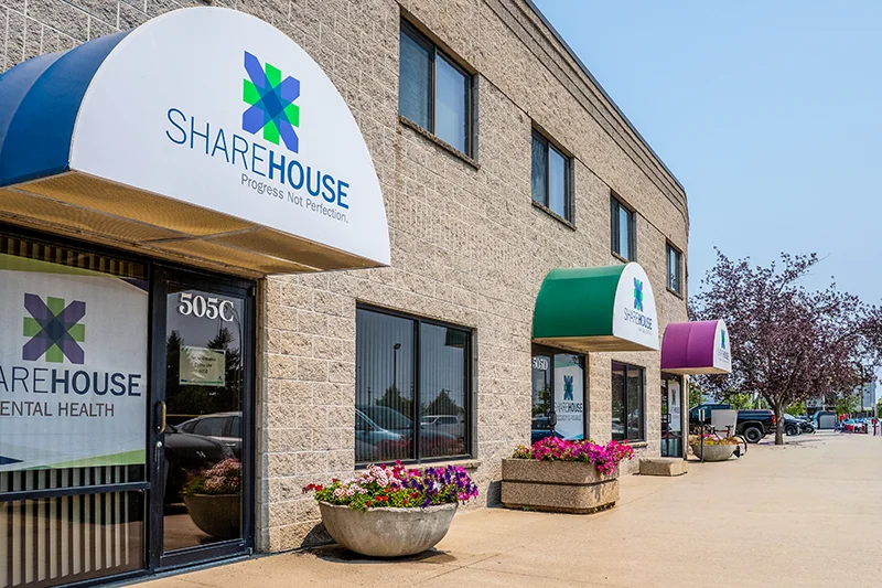 Exterior view of the ShareHouse Outpatient clinic building featuring three awnings, colorful flower planters, and a clear walkway leading to a parking area.