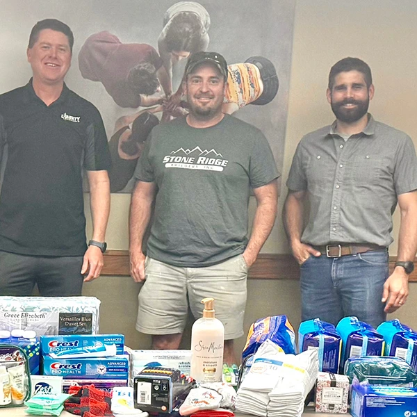 Three men standing at supply drive booth with essentials including shampoo, toothpaste and socks on the table.